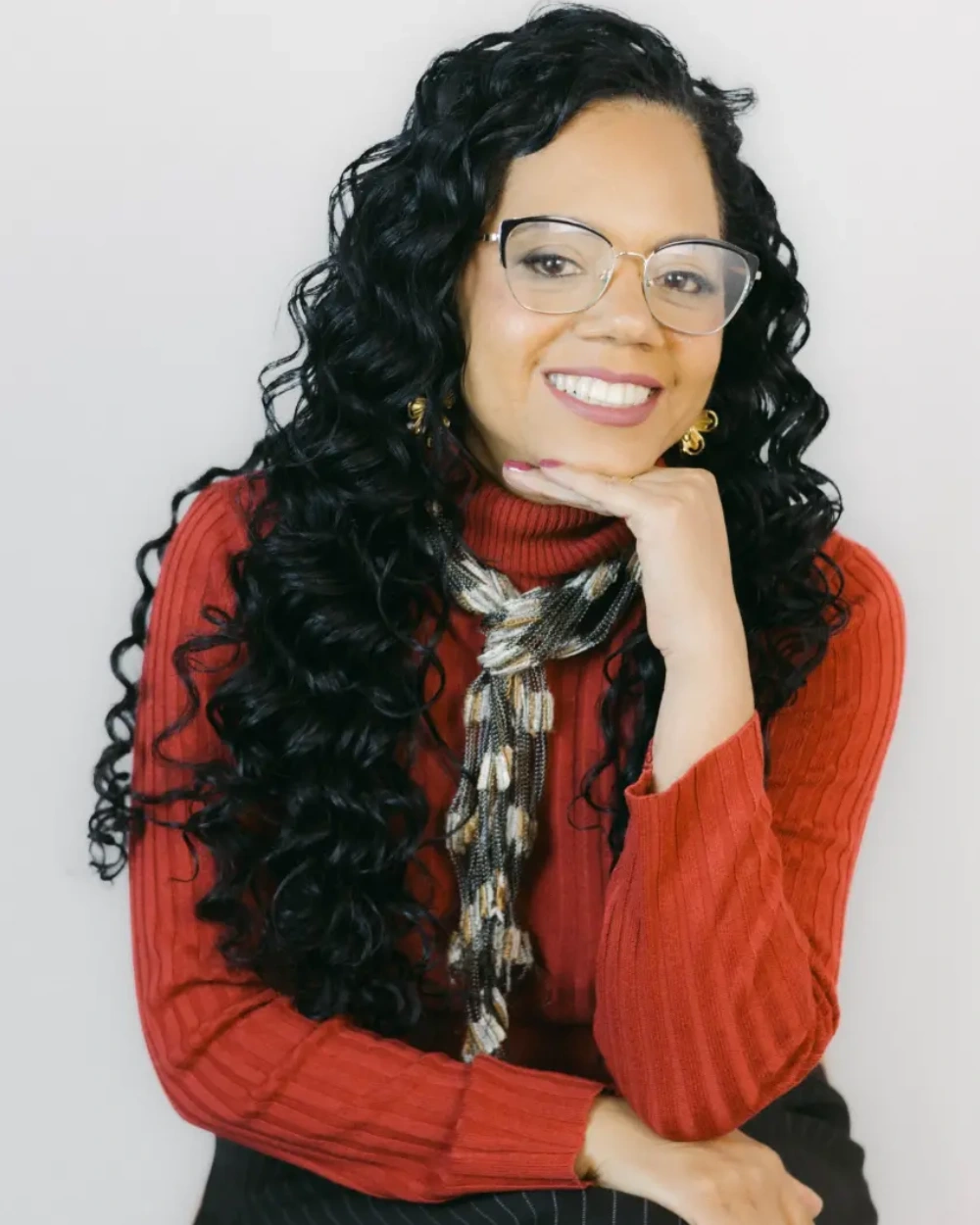 Roberta Vasconcelos de óculos e suéter vermelho, sorrindo com a mão apoiada no queixo